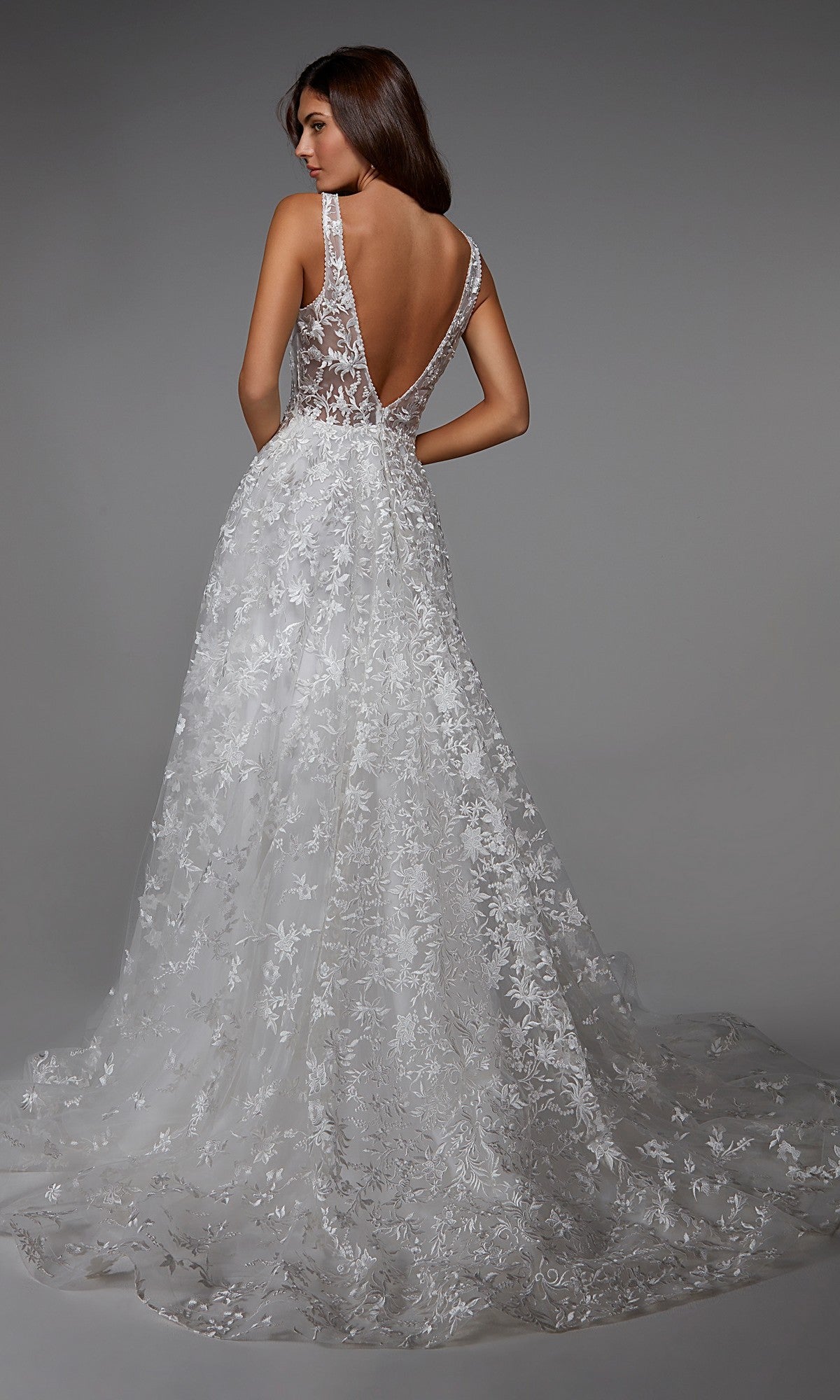 A woman wears the Long White Dress: Alyce 7037, a white sleeveless gown featuring a deep V-back and floral lace detailing, standing against a gray background.